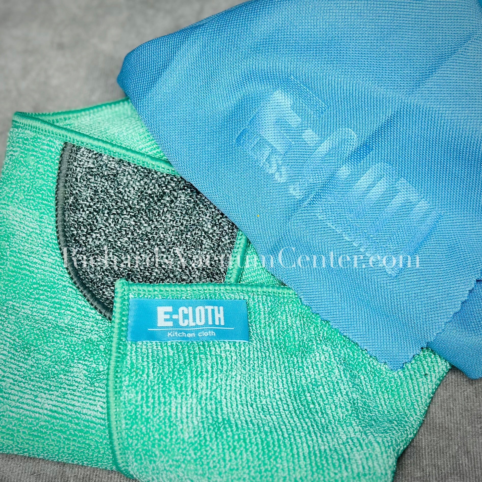 Two microfiber kitchen cleaning cloths, one green and one blue, with the E-Cloth brand visible on the green one, laid out on a grey surface.