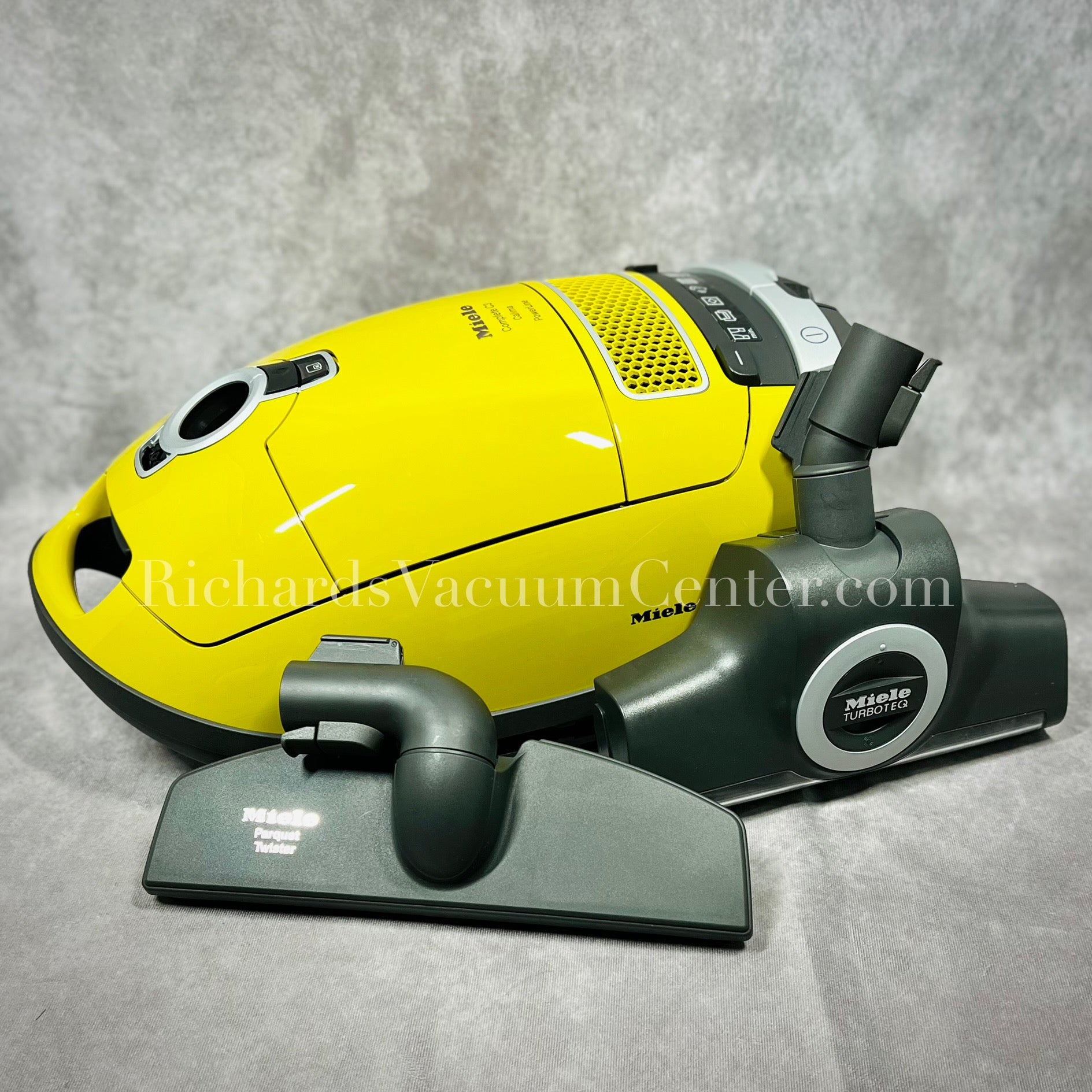A yellow Miele Complete C3 Calima vacuum cleaner with a gray floor brush attachment, photographed on a gray background.