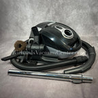 A black Titan T1400 deluxe canister vacuum with a telescopic wand, power cord, and premium accessory tools displayed.