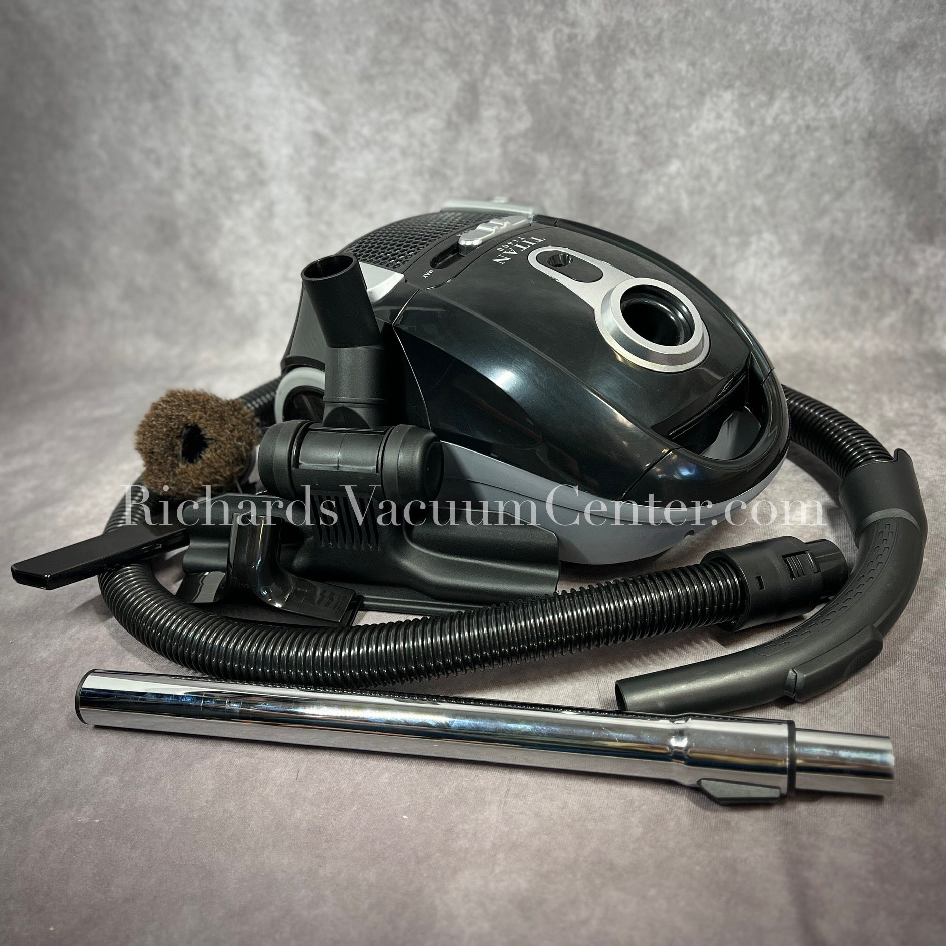 A black Titan T1400 deluxe canister vacuum with a telescopic wand, power cord, and premium accessory tools displayed.